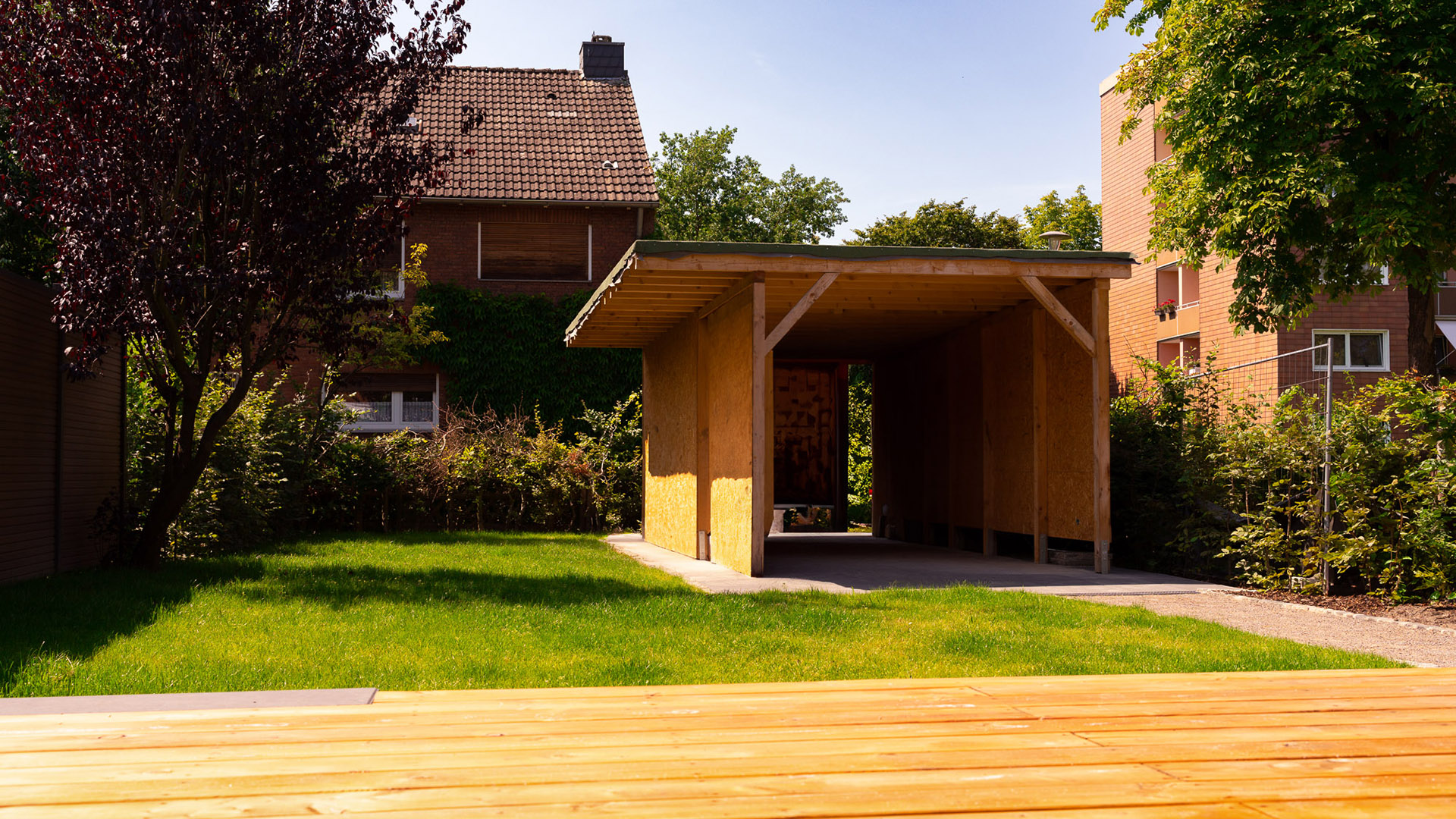 Holzschuppen im Garten mit grünem Rasen und umgebenden Bäumen bei sonnigem Wetter.