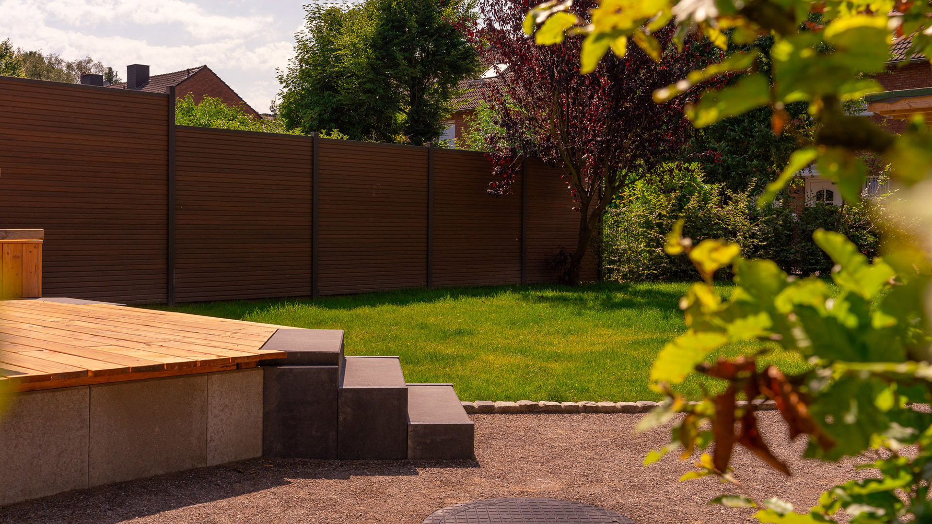 Garten mit Holzdeck und Treppe, umgeben von Rasen und Bäumen bei sonnigem Wetter.