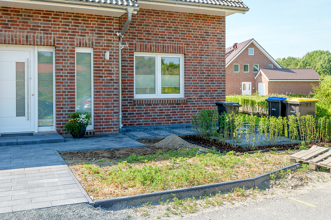 Ziegelhaus mit Garten. Grünfläche und gepflasterter Weg. Zwei Mülleimer stehen links.