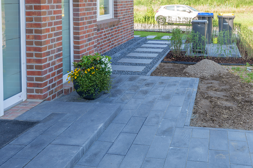 Moderner Hauseingang mit grauen Pflastersteinen, Backsteinfassade, Blumentopf und Mülltonnen.