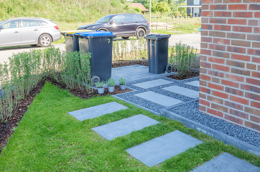 Ein moderner Gartenweg mit Pflastersteinen und Mülltonnen, gesäumt von grünen Pflanzen.