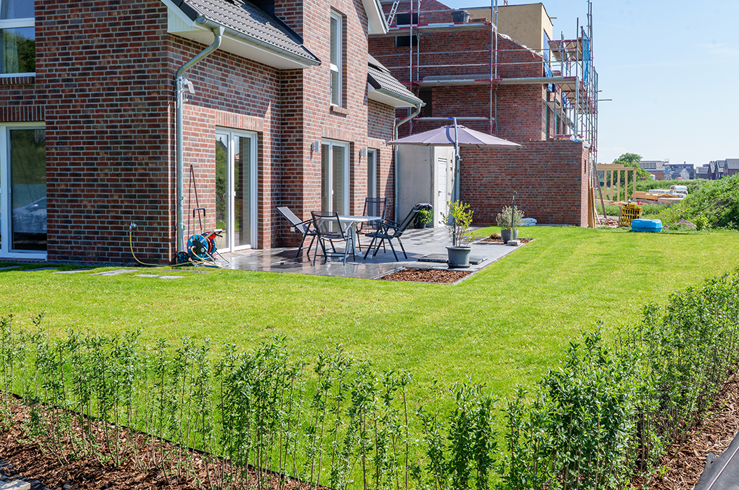 Moderne Backstein-Doppelhaushälfte mit gepflegtem Rasen, Terrasse und Baugerüst im Hintergrund.