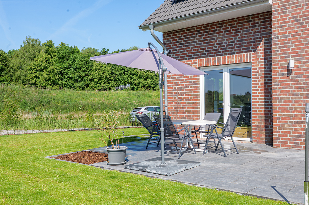 Eine Terrasse mit grauen Fliesen, Gartenmöbeln und violettem Sonnenschirm vor einem Backsteinhaus.