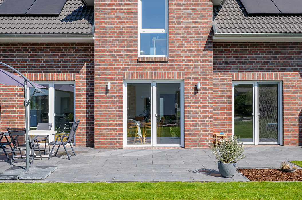Backstein-Haus mit grauen Dachziegeln, drei weißen Fenstertüren, Terrasse und Rasen im Vordergrund.