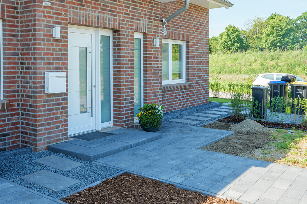 Backsteinfassade mit weißer Tür, Blumentopf, Pflastersteine, Kiesweg und Garten mit grünen Pflanzen.