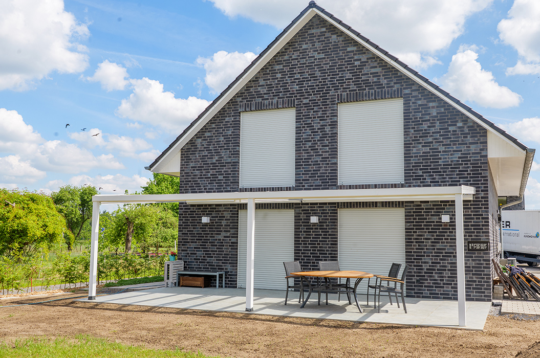 Neues Haus mit dunklen Ziegeln, weißem Terrassendach und Holzmöbeln im Garten bei sonnigem Himmel.