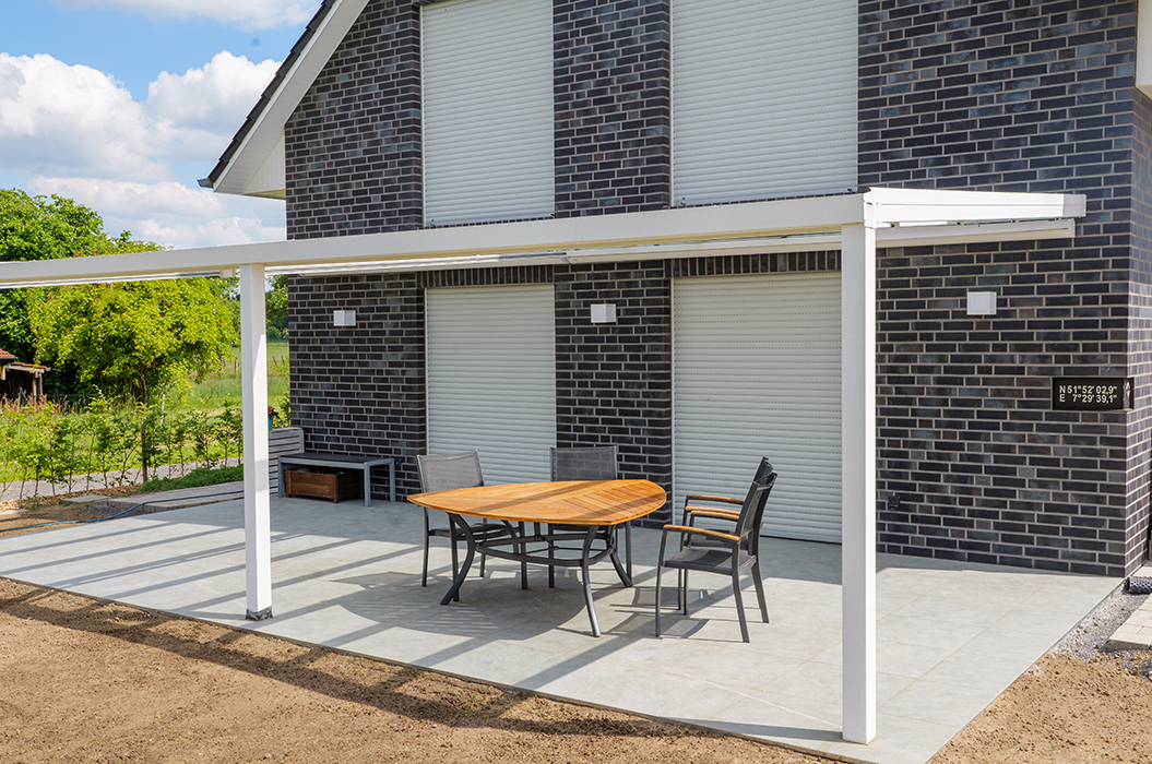 Ein moderner, grauer Ziegelbau mit Steinveranda und Holztisch unter weißem Pergola-Dach.