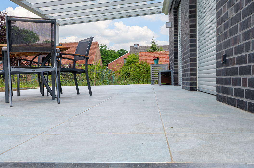 Moderne Terrasse mit grauem Bodenbelag, gläserner Überdachung und Tischgruppe vor Ziegelwand.
