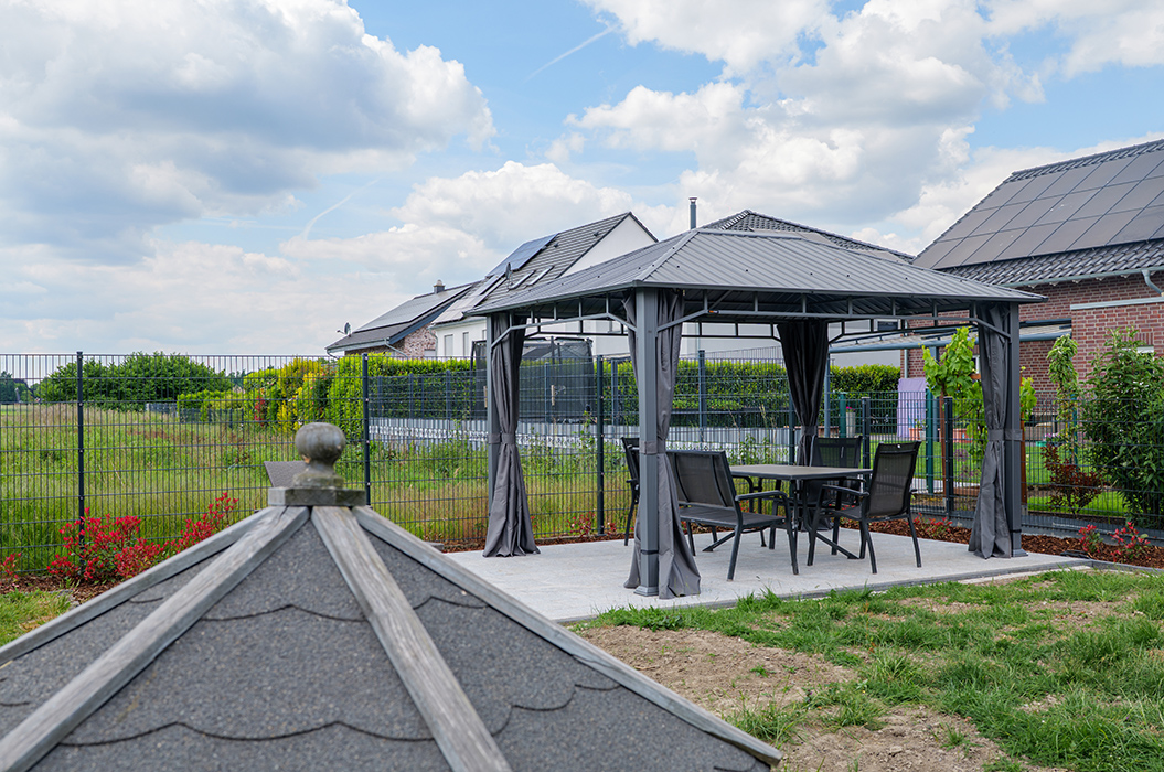 Ein grauer Pavillon mit Tisch und Stühlen in einem Garten mit Rasen und Wolken am Himmel.