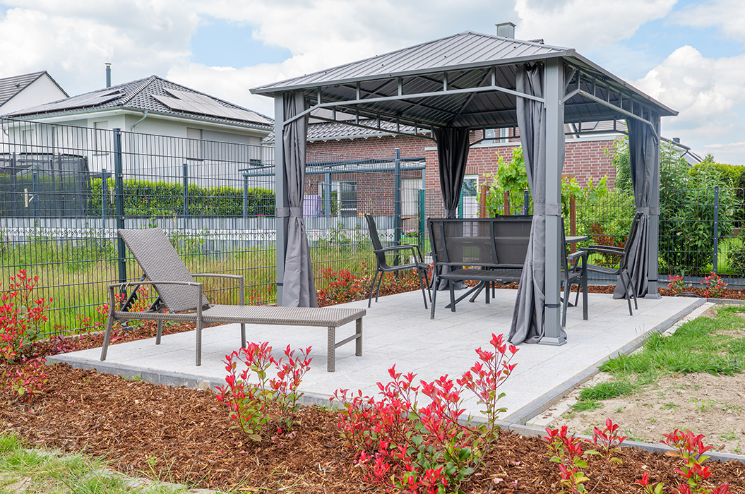 Pavillon im Garten mit grauen Vorhängen, Gartenmöbeln und roten Pflanzen im Vordergrund.