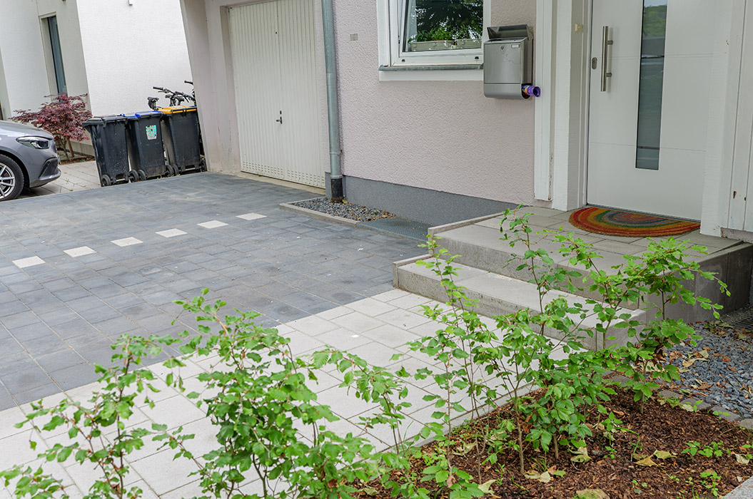Zufahrt und Eingang eines modernen Hauses mit grauen Pflastersteinen, Begrünung und Mülleimern.