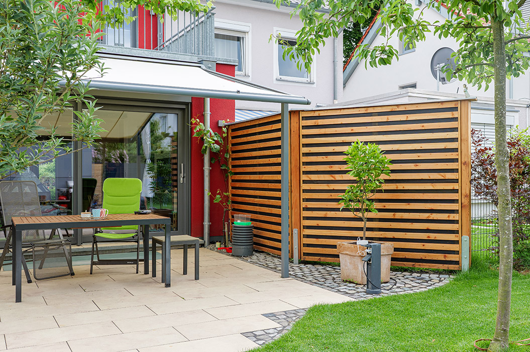 Eine sonnige Terrasse mit Tisch, grüner Stuhl, Holzsichtschutz und kleinen Bäumen.