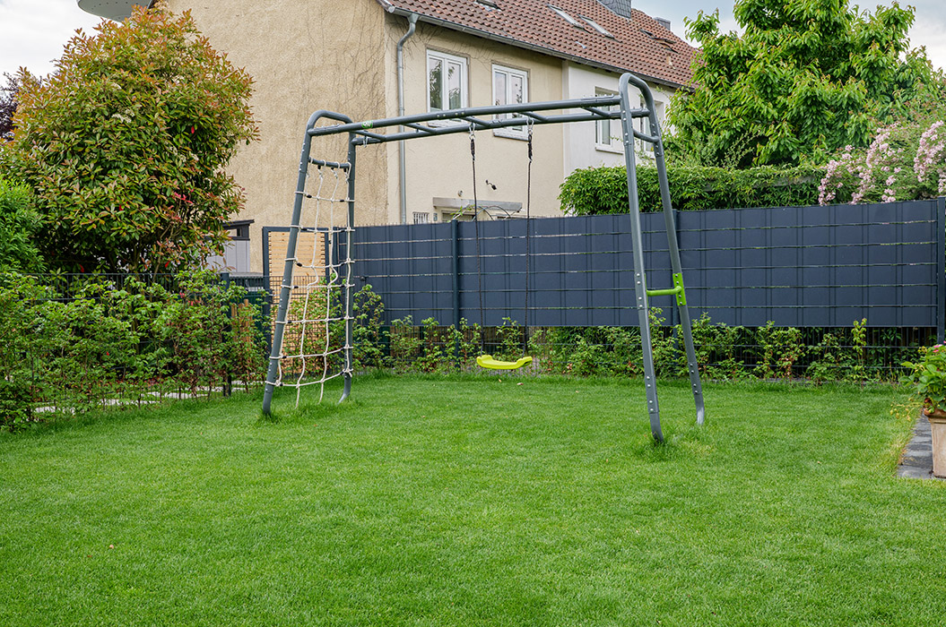 Schaukelgerüst im Garten mit Kletterwand, grünem Gras und Hintergrundhaus, umgeben von Büschen.
