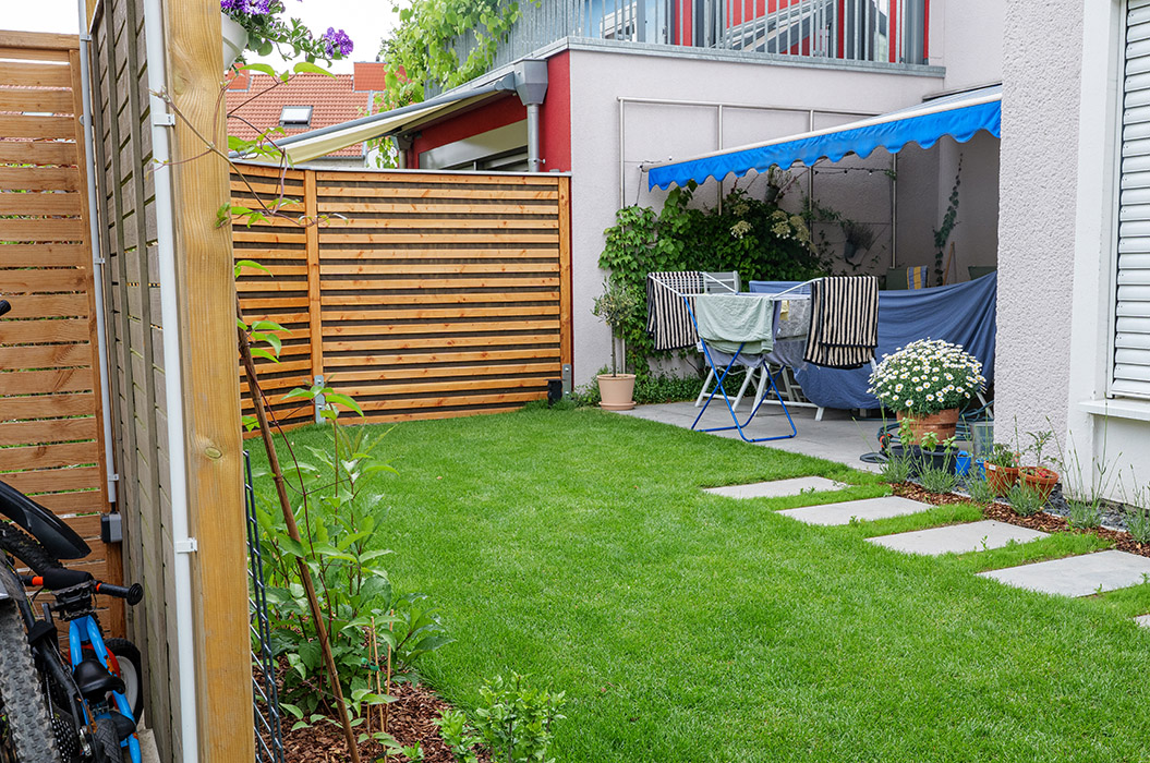 Ein gepflegter Garten mit Rasen, Terrasse, Tisch, Stühlen und blühenden Pflanzen im Hintergrund.