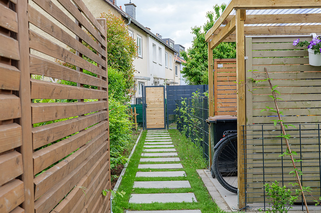 Ein schmaler, gepflasterter Gartenweg mit Holzzaun und kleiner Terrasse.