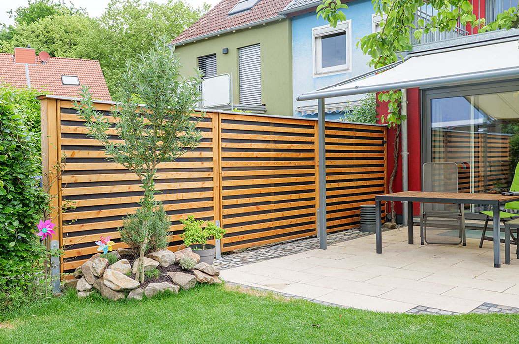 Gepflegter Garten mit Holz-Zaun, kleinen Bäumen, Terrasse und buntem Haus.