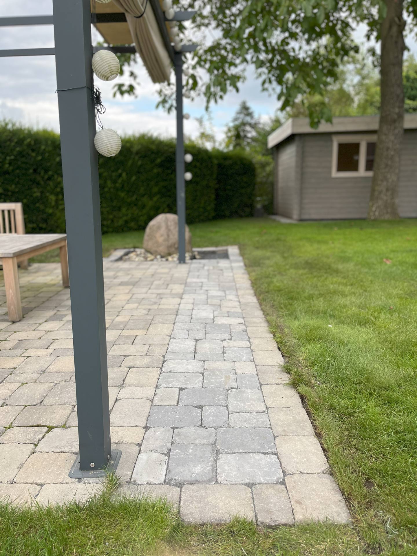 Ein gepflasterter Gartenbereich mit Holzbank neben grünem Rasen und Bäumen