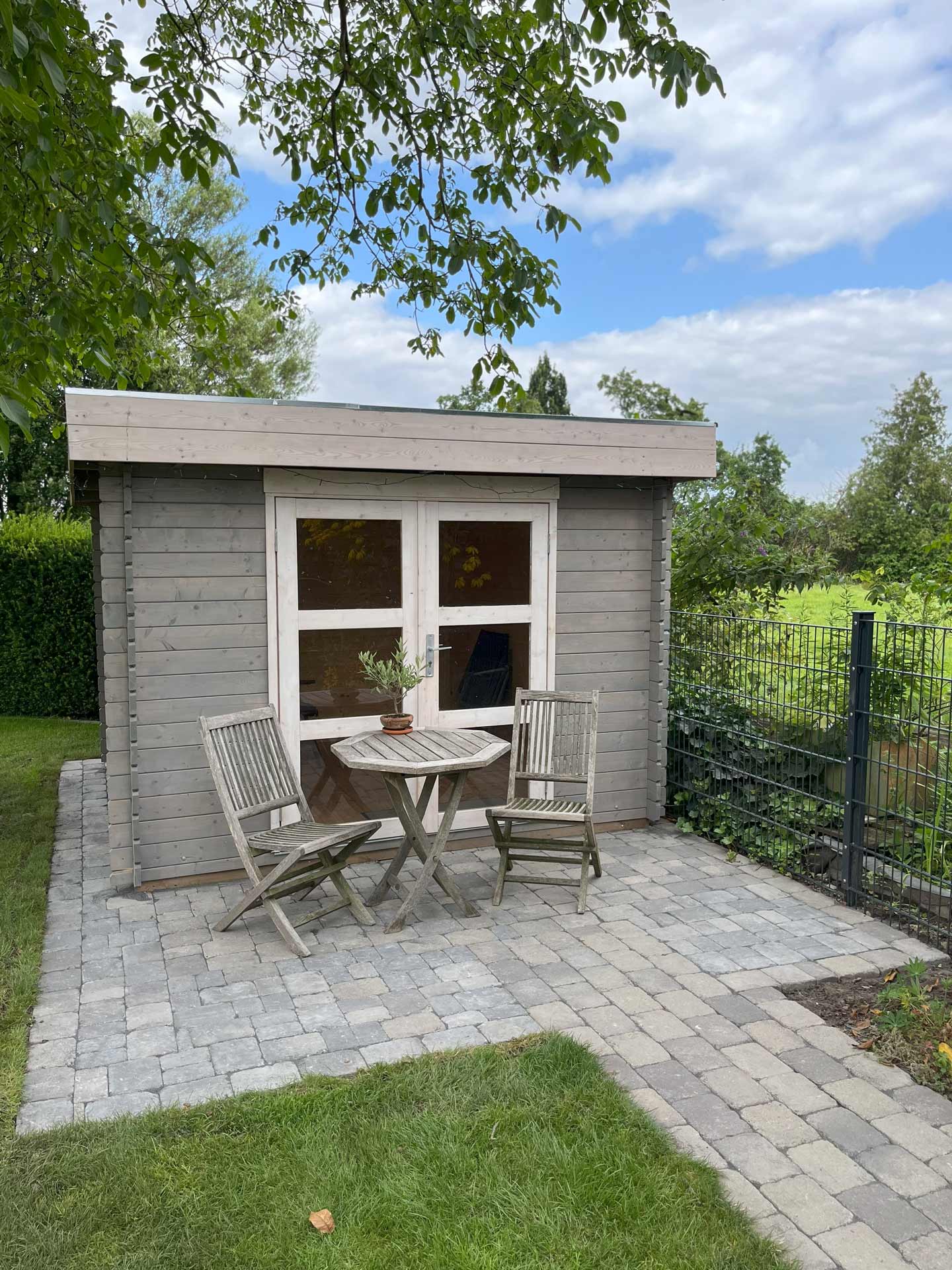 Holzschuppen mit grauem Anstrich, davor zwei Stühle und ein Tisch auf Steinterrasse.