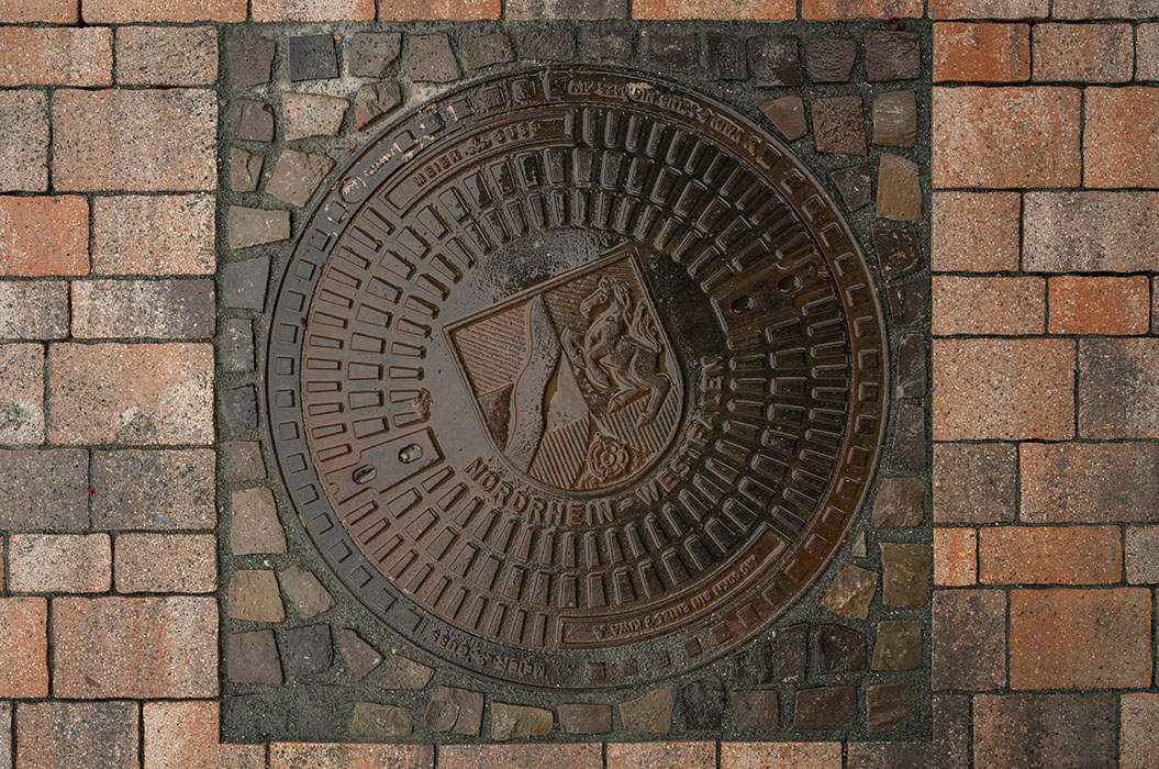 Ein runder Kanaldeckel aus Metall mit Wappen von Nordrhein-Westfalen auf rötlichen Pflastersteinen