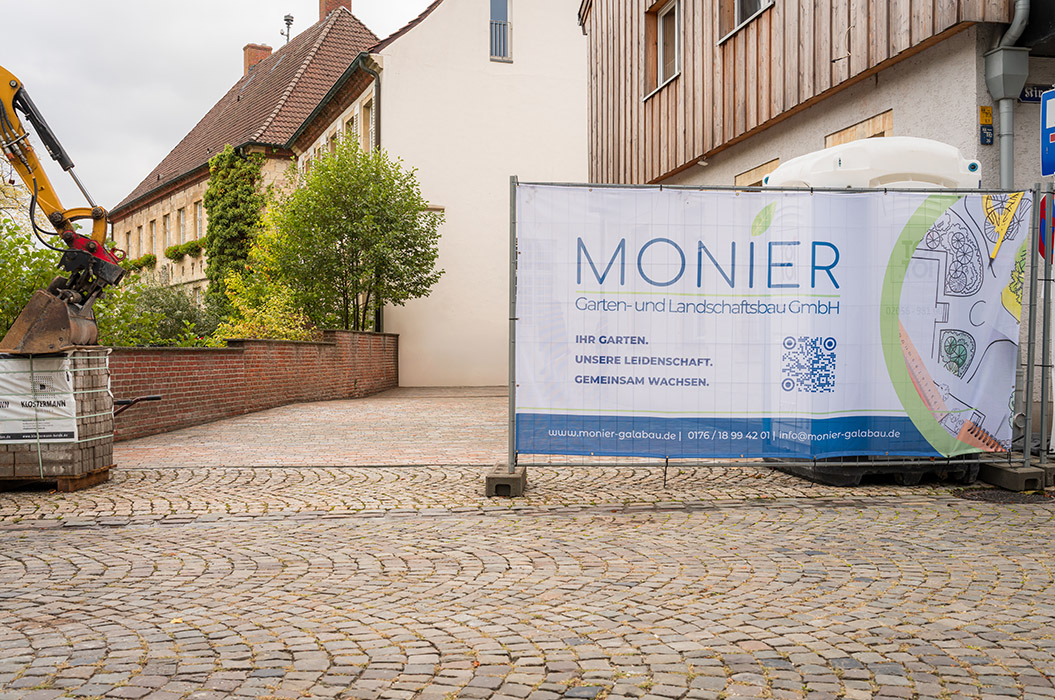 Bauzaun mit MONIER Werbeplane vor Kopfsteinpflaster und alten Gebäuden