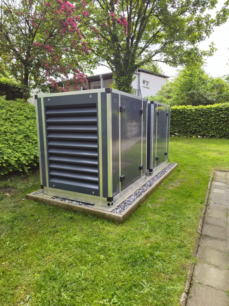 Grau gruene Technikbox mit Lamellenfront im Garten neben Hecke und Baum