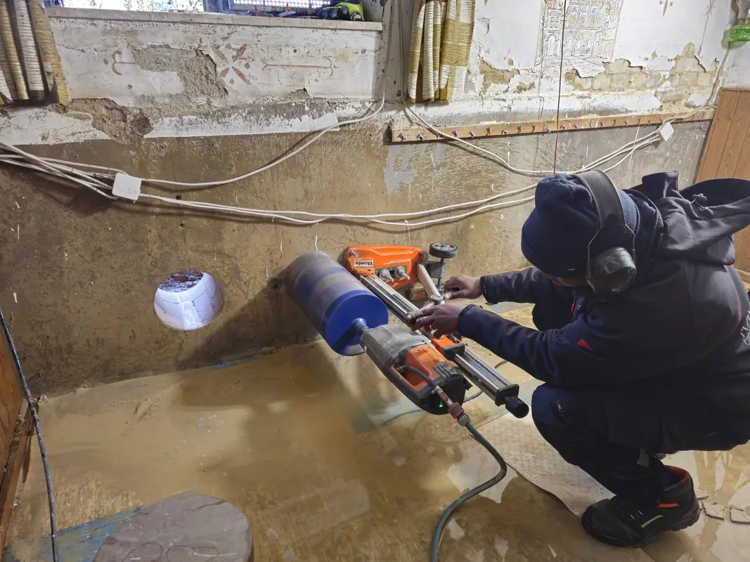 Kauernder Arbeiter mit Gehoerschutz bohrt nassen Beton mit orangeblauer Bohrmaschine