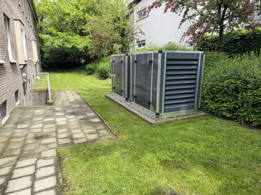 Graue Technikcontainer mit Lüftungsgitter auf Grünfläche neben Pflasterweg, still und markant