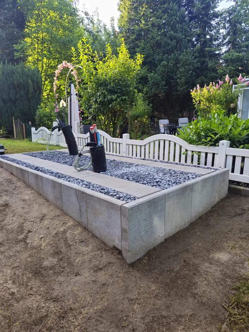 Grauer Kieskasten mit zwei schwarzen Pumpen vor weißem Zaun im Garten