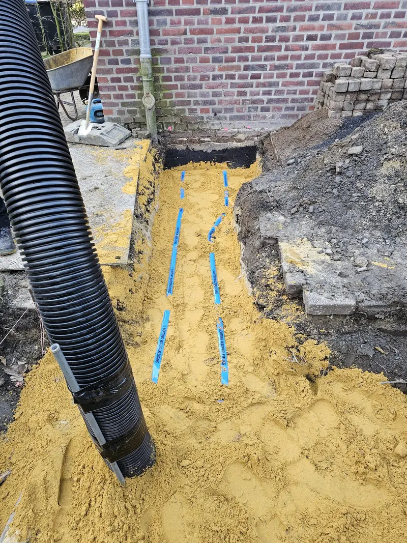 Sandgrube mit blauen Leitungen, schwarzem Rohr und Ziegelwand im Hintergrund