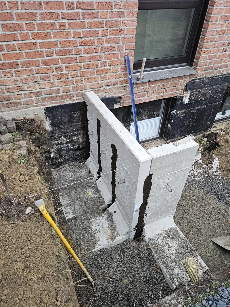 Graue Betonmauer vor roter Backsteinwand, Baustelle mit Wasserwaage und Kies
