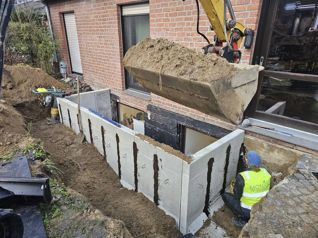 Bauarbeiter mit neutralem Blick im gelben Helm am Erdschacht mit Bagger und Sand