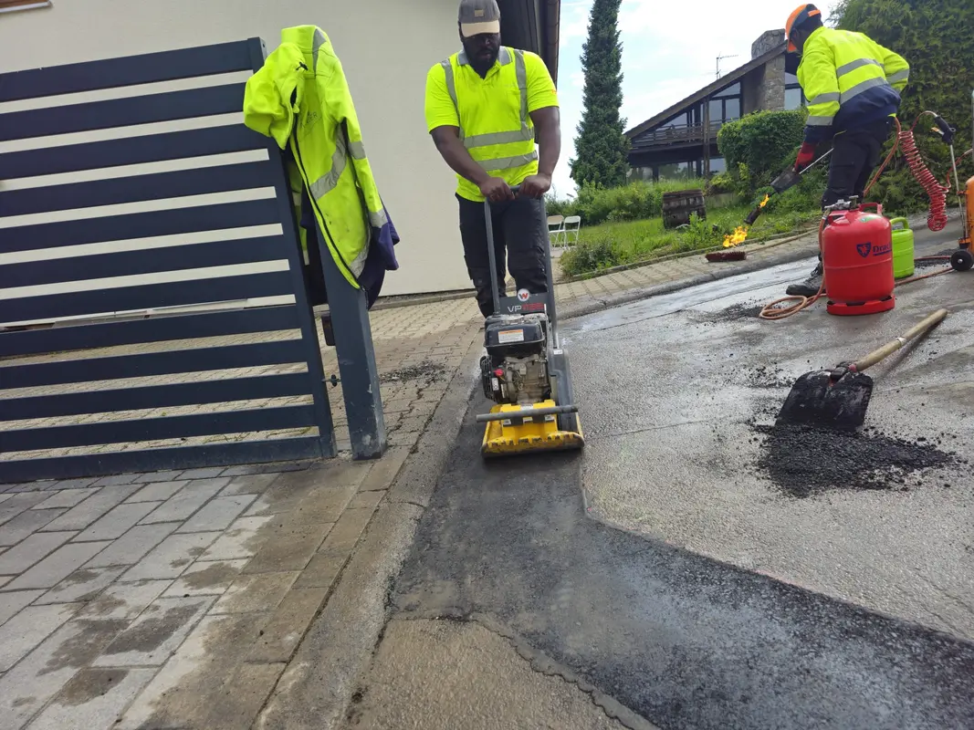 Zwei Männer in gelben Westen arbeiten konzentriert an frischem Asphalt, mit Rüttler und Brenner