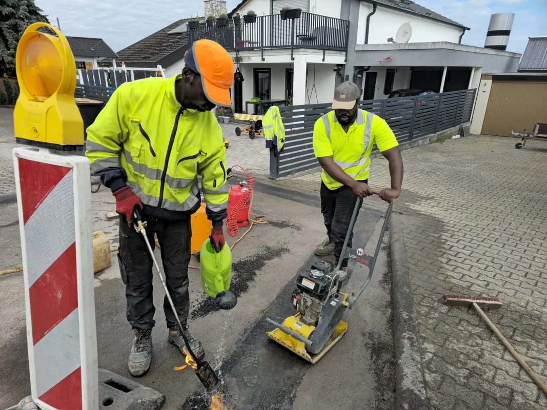 Zwei Bauarbeiter konzentriert, gelbe Jacken, Asphaltarbeiten mit Maschine und Schlauch